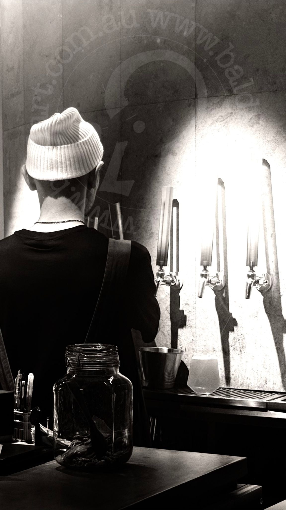 Person wearing a beanie standing in front of a bar with glasses and bottles.