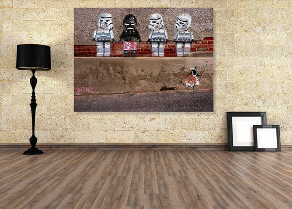 Framed artwork of four stormtroopers and a penguin on a brick wall, displayed on a wall with a lamp and picture frames.