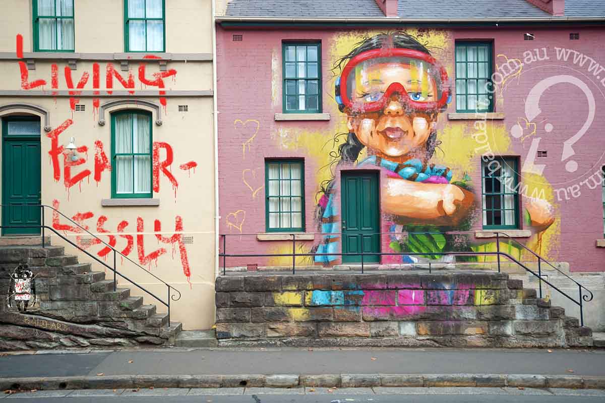 A masked woman poses near a building adorned with vibrant graffiti, showcasing street art in an urban environment.