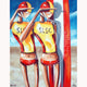 A painting featuring two stylized figures in yellow and red swimwear against a beach and surf-themed background, signed by the artist.